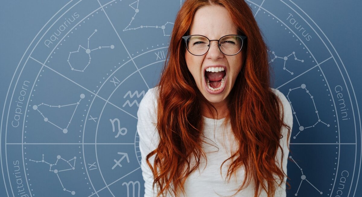 Eine rothaarige Frau mit Brille schreit wütend oder frustriert in die Kamera. Im Hintergrund ist ein astrologischer Kreis mit den Sternzeichen-Symbolen auf einem blauen Hintergrund zu sehen.
