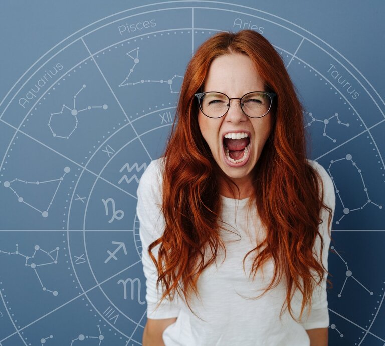 Eine rothaarige Frau mit Brille schreit wütend oder frustriert in die Kamera. Im Hintergrund ist ein astrologischer Kreis mit den Sternzeichen-Symbolen auf einem blauen Hintergrund zu sehen. | © Getty Images/ stockfour und WinWin.artlab via Canva.com [M]