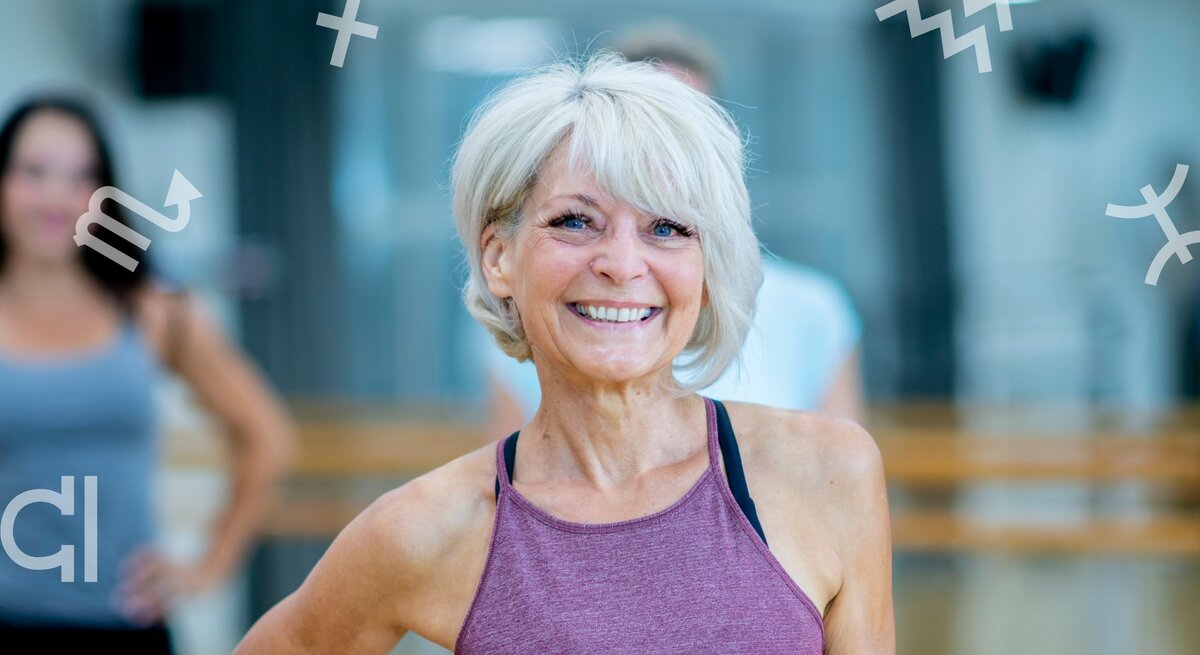 Eine ältere, sportliche Frau mit grauem Haar und lila Sporttop lächelt selbstbewusst in die Kamera. Sie befindet sich in einem hellen Fitnessstudio. Im Hintergrund sind unscharf weitere Personen zu sehen. Um sie herum schweben verschiedene astrologische Sternzeichen-Symbole, was auf einen Zusammenhang zwischen Fitness und Astrologie hinweist.