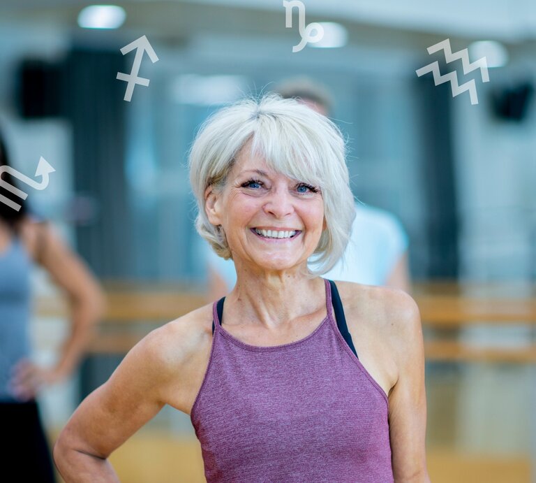 Eine ältere, sportliche Frau mit grauem Haar und lila Sporttop lächelt selbstbewusst in die Kamera. Sie befindet sich in einem hellen Fitnessstudio. Im Hintergrund sind unscharf weitere Personen zu sehen. Um sie herum schweben verschiedene astrologische Sternzeichen-Symbole, was auf einen Zusammenhang zwischen Fitness und Astrologie hinweist. | © Getty Images Signature/ FatCamera und Chikovnaya via Canva.com [M]