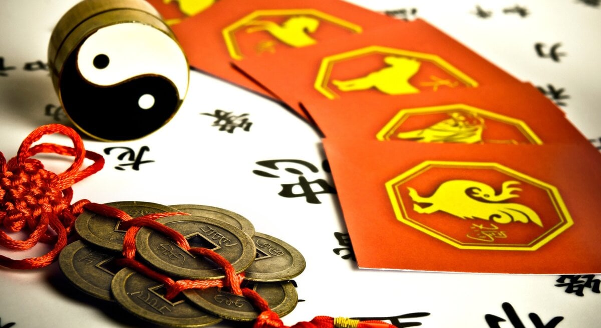  Eine detailreiche Darstellung chinesischer Astrologie: Im Vordergrund liegt ein traditioneller Glücksbringer mit Münzen und rotem Band. Daneben steht ein Yin-Yang-Symbol, umgeben von roten Karten mit gelben Illustrationen der chinesischen Tierkreiszeichen. Der Hintergrund zeigt schwarze chinesische Schriftzeichen auf weißem Papier. Das Bild vermittelt eine Atmosphäre von Tradition, Weisheit und fernöstlicher Kultur.