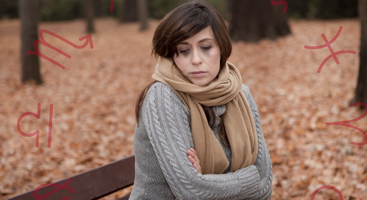 Eine nachdenkliche Frau sitzt mit verschränkten Armen auf einer Parkbank an einem Herbsttag. Sie trägt einen grauen Strickpullover und einen beigen Schal. Um sie herum liegen gefallene Blätter. Im Hintergrund sind Symbole von Sternzeichen zu sehen, die in Rot über das Bild gelegt sind. Die Frau wirkt bedrückt oder nachdenklich, möglicherweise in Verbindung mit einer Herbstdepression.