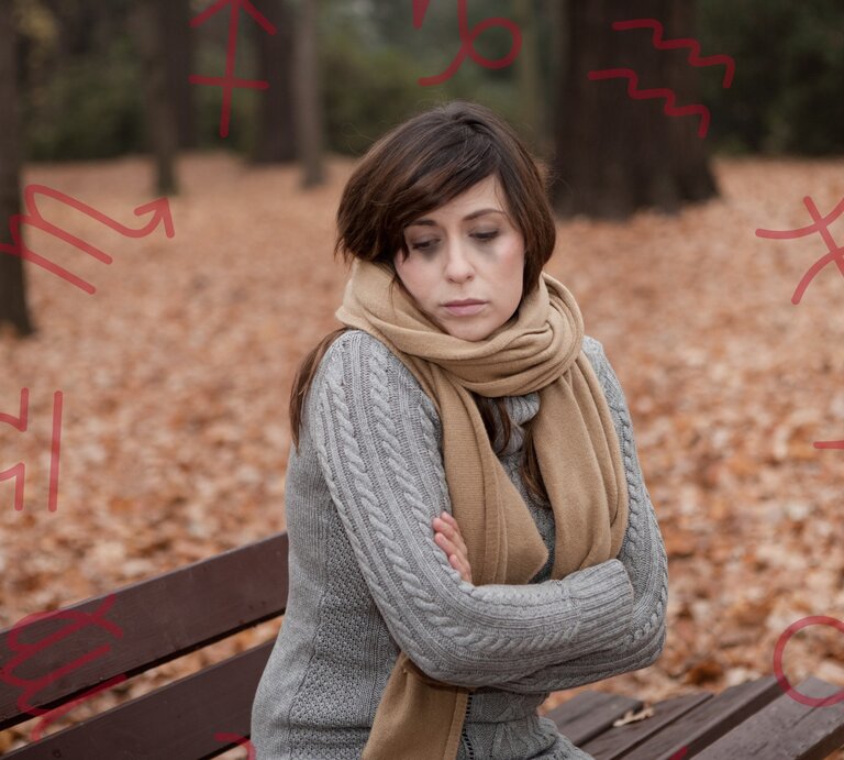 Eine nachdenkliche Frau sitzt mit verschränkten Armen auf einer Parkbank an einem Herbsttag. Sie trägt einen grauen Strickpullover und einen beigen Schal. Um sie herum liegen gefallene Blätter. Im Hintergrund sind Symbole von Sternzeichen zu sehen, die in Rot über das Bild gelegt sind. Die Frau wirkt bedrückt oder nachdenklich, möglicherweise in Verbindung mit einer Herbstdepression. | © Adobe Stock/ drubig-photo und Keronyart's Images/ Be keronyart via Canva.com [M]