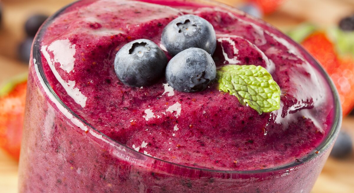 Ein dichter, lila-roter Smoothie in einem Glas, garniert mit frischen Blaubeeren und einem Minzblatt. Im Hintergrund sind verschwommene Erdbeeren und Blaubeeren zu sehen.