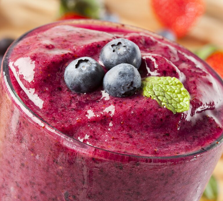 Ein dichter, lila-roter Smoothie in einem Glas, garniert mit frischen Blaubeeren und einem Minzblatt. Im Hintergrund sind verschwommene Erdbeeren und Blaubeeren zu sehen. | © pixabay/ dennisyang988 via Canva.com