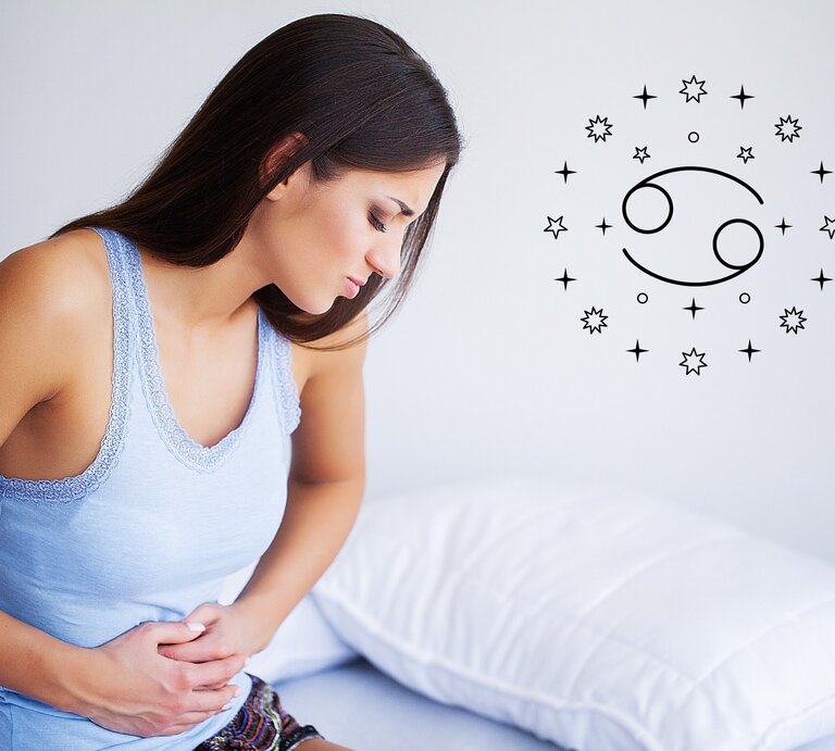 Das Bild zeigt eine junge Frau mit langen braunen Haaren, die ein hellblaues Oberteil trägt und sich vor Schmerzen den Bauch hält. Sie sitzt auf einem Bett mit einem weißen Kissen und einem weißen Bettlaken. Neben ihr ist das astrologische Symbol für Krebs, umgeben von kleinen Sternen, zu sehen. Die Frau wirkt angespannt und unwohl, was auf Bauchschmerzen oder Verdauungsprobleme hinweist. Das astrologische Symbol deutet darauf hin, dass das Bild in einem Zusammenhang mit einem Gesundheits-Horoskop für das Sternzeichen Krebs steht. | © Iurii Maksymiv und warmjuly via Canva.com [M]