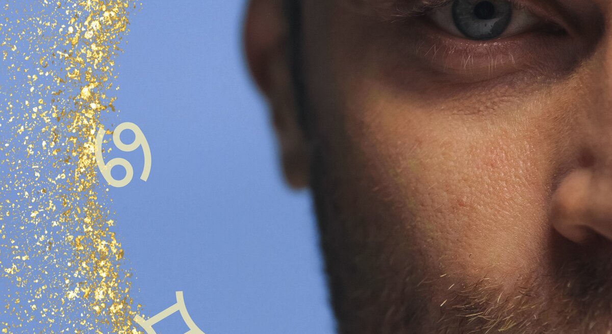 Nahaufnahme eines Mannes, dessen Gesicht nur zur Hälfte zu sehen ist. Der Fokus liegt auf seinem blauen Auge und einem Teil seines bärtigen Gesichts. Der Hintergrund ist hellblau und mit goldenen Glitzerpartikeln verziert. Um das Bild herum sind verschiedene Sternzeichen-Symbole verteilt, was eine astrologische Thematik andeutet. Die Komposition vermittelt eine geheimnisvolle und nachdenkliche Atmosphäre.