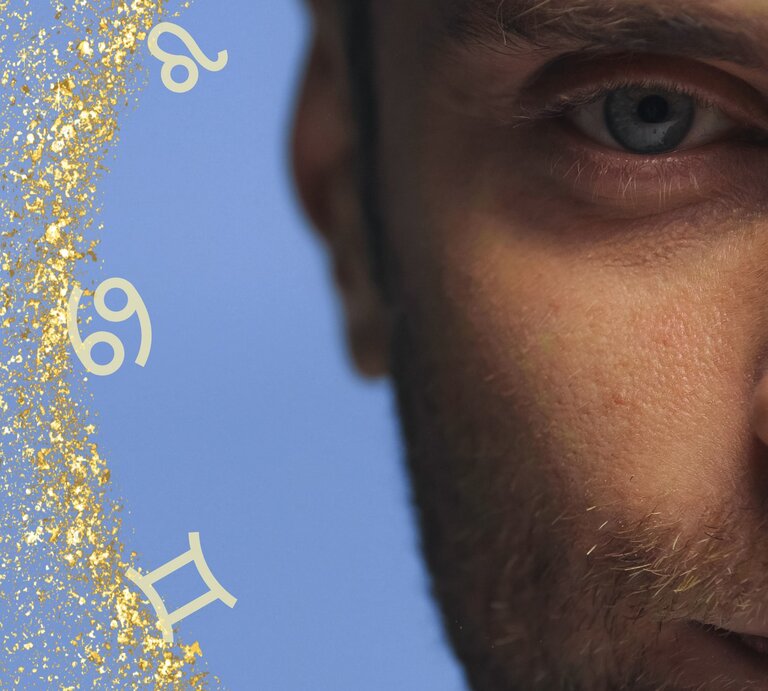 Nahaufnahme eines Mannes, dessen Gesicht nur zur Hälfte zu sehen ist. Der Fokus liegt auf seinem blauen Auge und einem Teil seines bärtigen Gesichts. Der Hintergrund ist hellblau und mit goldenen Glitzerpartikeln verziert. Um das Bild herum sind verschiedene Sternzeichen-Symbole verteilt, was eine astrologische Thematik andeutet. Die Komposition vermittelt eine geheimnisvolle und nachdenkliche Atmosphäre. | © Adobe Stock/ fotoart89 (KI-generiert), JMBee Studio und Chikovnaya via Canva.com [M]