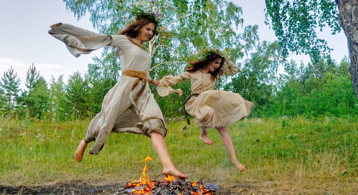 Zwei Frauen mit Blumenkränzen springen über ein Sonnenwend-Feuer.