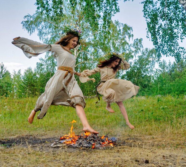Zwei Frauen mit Blumenkränzen springen über ein Sonnenwend-Feuer. | © Adobe Stock/ Aikon