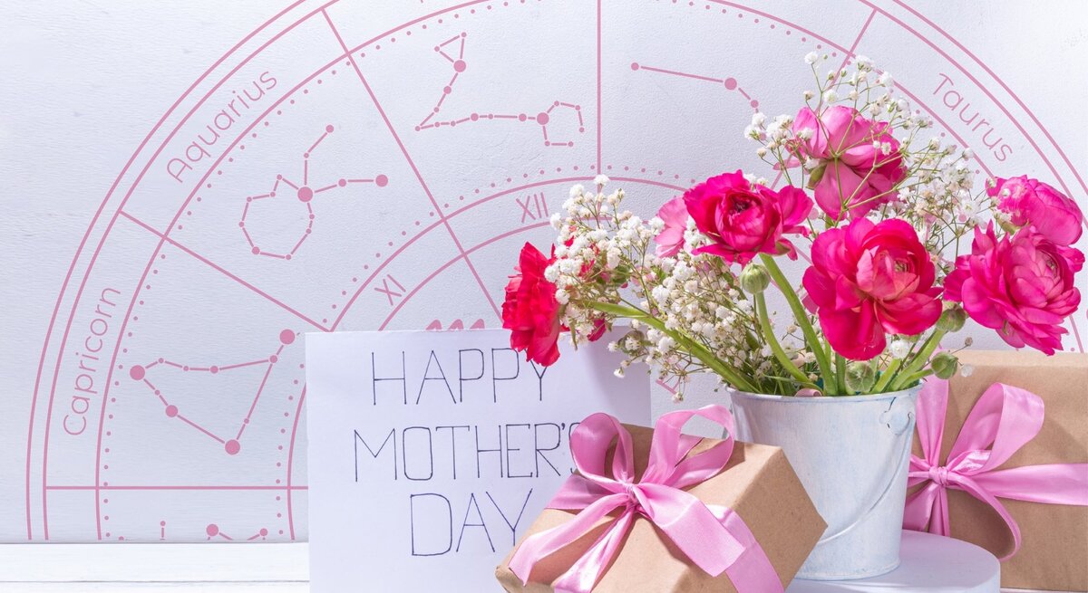 Ein liebevoll arrangiertes Muttertagsstillleben mit pinken Ranunkeln und Schleierkraut in einem weißen Metalltopf, daneben zwei in braunes Papier eingepackte Geschenke mit rosa Schleifen und eine Karte mit der Aufschrift ‚Happy Mother’s Day‘. Im Hintergrund ist ein rosafarbener Tierkreis mit Sternzeichen wie Pisces, Taurus und Capricorn zu sehen – eine harmonische Verbindung aus Astrologie, Blumen und Muttertagsliebe.