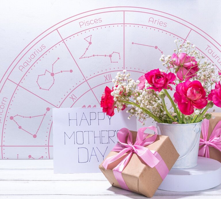 Ein liebevoll arrangiertes Muttertagsstillleben mit pinken Ranunkeln und Schleierkraut in einem weißen Metalltopf, daneben zwei in braunes Papier eingepackte Geschenke mit rosa Schleifen und eine Karte mit der Aufschrift ‚Happy Mother’s Day‘. Im Hintergrund ist ein rosafarbener Tierkreis mit Sternzeichen wie Pisces, Taurus und Capricorn zu sehen – eine harmonische Verbindung aus Astrologie, Blumen und Muttertagsliebe. | © rimmabondarenko und WinWin.artlab via Canva.com [M]