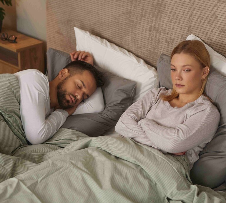 Verärgerte Frau denkt neben ihrem schlafenden Partner über Beziehungsprobleme und die Gleichgültigkeit des Partners nach. | © stockphotodirectors/Getty Images