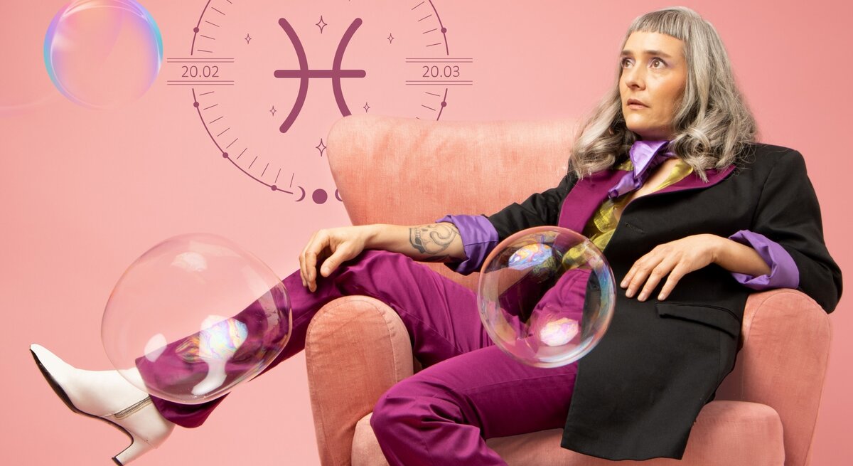 Eine Frau mit langen grauen Haaren trägt einen lila Retro-Anzug und sitzt in einem Sessel, das Bein lässig über die Lehne geschlagen. Sie ist umgeben von Seifenblasen und neben ihr ist das astrologische Symbol des Sternzeichens Fische in einem Kreis zu sehen.