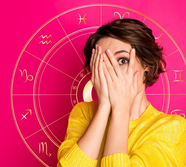 Eine überraschte Frau in einem gelben Oberteil hält ihre Hände vor ihr Gesicht und späht durch ihre Finger. Im Hintergrund ist eine leuchtend rosa Grafik mit astrologischen Symbolen, darunter das Rad des Tierkreises mit den Zeichen von Widder bis Fische. | © Adobe Stock/ deagreez und Artulina via Canva.com [M]