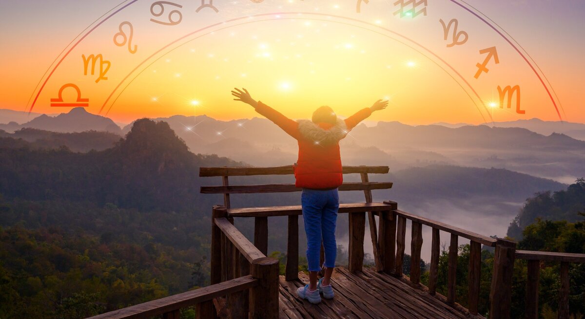 Ein Mensch steht mit ausgebreiteten Armen auf einer Holzplattform und bewundert den Sonnenaufgang über einem bergigen Landschaftspanorama. Über dem Bild ist ein halbkreisförmiger Bogen mit astrologischen Symbolen der Tierkreiszeichen von Wassermann bis Steinbock gezeichnet, die in einem Farbverlauf von Gelb zu Rot angeordnet sind, passend zur aufgehenden Sonne im Hintergrund.