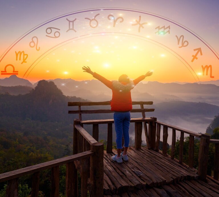 Ein Mensch steht mit ausgebreiteten Armen auf einer Holzplattform und bewundert den Sonnenaufgang über einem bergigen Landschaftspanorama. Über dem Bild ist ein halbkreisförmiger Bogen mit astrologischen Symbolen der Tierkreiszeichen von Wassermann bis Steinbock gezeichnet, die in einem Farbverlauf von Gelb zu Rot angeordnet sind, passend zur aufgehenden Sonne im Hintergrund. | © Adobe Stock/ sarayut_sy