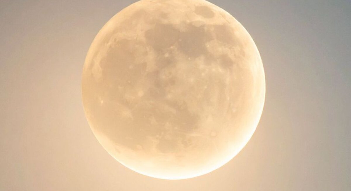 Ein Vollmond am Nachthimmel mit sanftem, goldenem Schimmer. Die Oberflächenstrukturen des Mondes, wie Krater und Ebenen, sind klar erkennbar. Der Himmel wirkt leicht aufgehellt und hat nur einen dezenten Blauton.