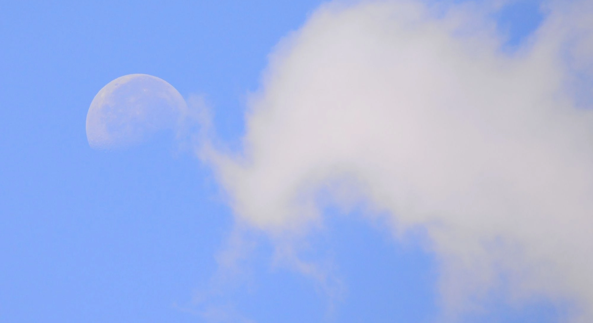 Halbmond am hellblauen Himmel, teilweise von einer weißen Wolke verdeckt.