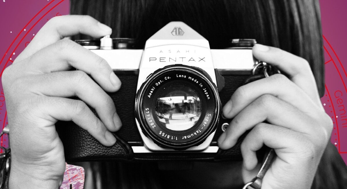 Eine Person hält eine analoge Kamera vom Typ Asahi Pentax vor das Gesicht, bereit zum Fotografieren. Im Hintergrund ist ein stilisiertes astrologisches Rad mit Sternzeichen auf violettem Grund zu sehen – Symbol für den Blick in die Zukunft und das Zusammenspiel von Kreativität und kosmischem Einfluss.