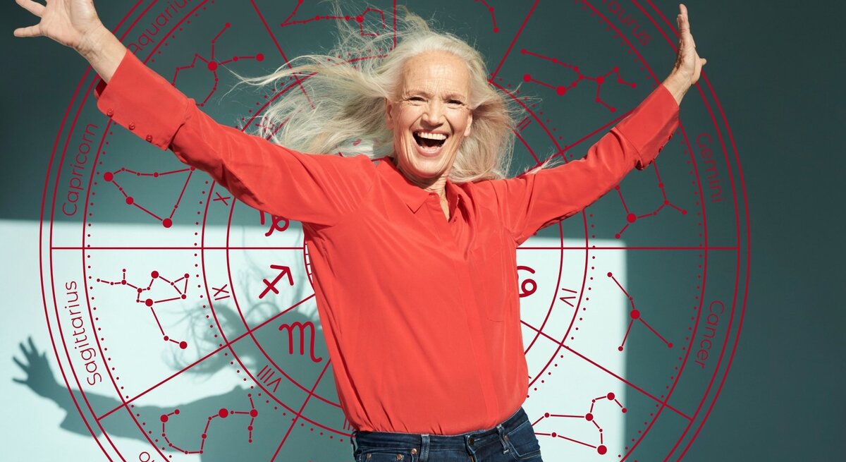 Eine ältere Frau mit langen grauen Haaren trägt ein rotes Oberteil und lacht mit ausgebreiteten Armen in die Kamera. Hinter ihr ist ein großes Horoskop-Rad mit den zwölf Sternzeichen und ihren Sternbildern zu sehen. Symbolische Darstellung von Lebensfreude und Glück im Zusammenhang mit Sternzeichen.