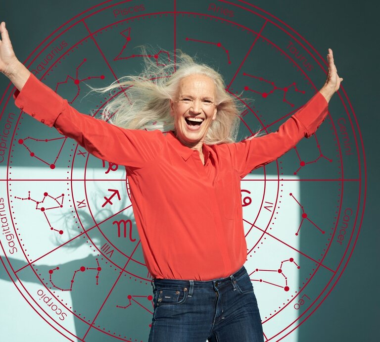 Eine ältere Frau mit langen grauen Haaren trägt ein rotes Oberteil und lacht mit ausgebreiteten Armen in die Kamera. Hinter ihr ist ein großes Horoskop-Rad mit den zwölf Sternzeichen und ihren Sternbildern zu sehen. Symbolische Darstellung von Lebensfreude und Glück im Zusammenhang mit Sternzeichen. | © Getty Images/ Flashpop und WinWin.artlab via Canva.com [M]