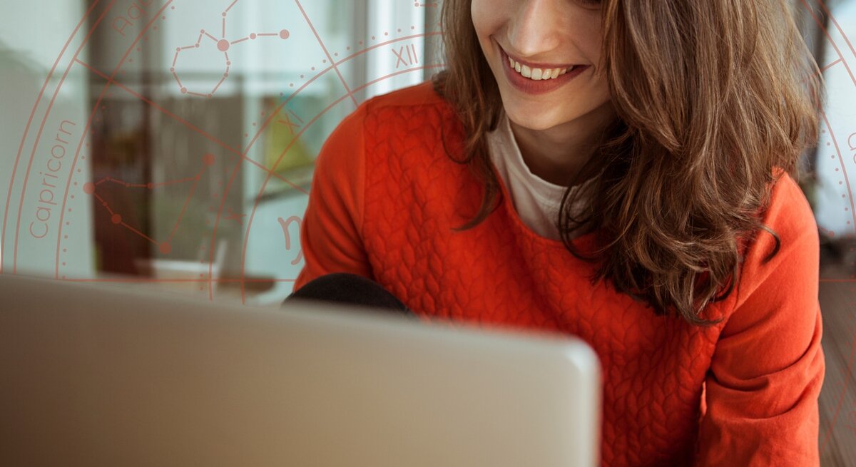 Eine lächelnde Frau mit schulterlangem braunen Haar sitzt an einem Tisch und schaut auf ihren Laptop-Bildschirm. Sie trägt einen roten Pullover. Im Hintergrund ist eine dezente, transparente Grafik eines Horoskop-Kreises mit Sternzeichen und Konstellationen eingeblendet.