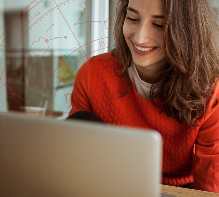 Eine lächelnde Frau mit schulterlangem braunen Haar sitzt an einem Tisch und schaut auf ihren Laptop-Bildschirm. Sie trägt einen roten Pullover. Im Hintergrund ist eine dezente, transparente Grafik eines Horoskop-Kreises mit Sternzeichen und Konstellationen eingeblendet. | © Getty Images/ Westend61 und WinWin.artlab via Canva.com [M]