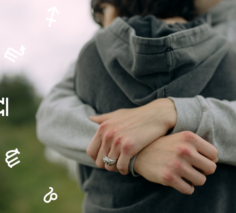 Ein Paar umarmt sich innig im Freien, beide tragen graue Hoodies. Die Hände der Personen sind sichtbar, mit leicht geröteten Knöcheln. Im Hintergrund sind unscharfe Bäume oder Natur zu erkennen. Um das Paar schweben astrologische Symbole verschiedener Sternzeichen, was dem Bild eine mystische, horoskopbezogene Atmosphäre verleiht. | © Pexels/ cottonbro studio und Winwin.artlab via Canva.com [M]