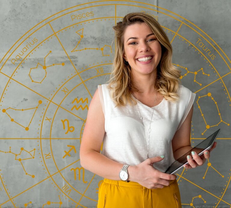 Eine junge Frau mit schulterlangem, welligem blondem Haar steht lächelnd vor einer grauen Wand mit einem goldenen astrologischen Tierkreis. Sie trägt eine weiße Bluse, eine gelbe Hose und eine silberne Uhr am Handgelenk. In ihren Händen hält sie ein Tablet. Der Hintergrund zeigt die Sternzeichen mit ihren Symbolen und Sternbildern in einem kreisförmigen Design. | © Getty Images/ StockRocket und WinWin.artlab via Canva.com