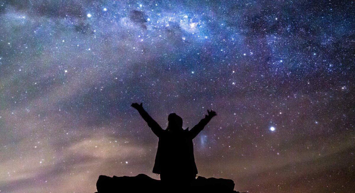 Eine Silhouette einer Person mit ausgestreckten Armen steht auf einem Felsen unter einem beeindruckenden Sternenhimmel. Die Milchstraße leuchtet hell über ihr, während der Himmel in tiefen Blau- und Violetttönen schimmert. Die Szene strahlt ein Gefühl von Freiheit, Staunen und Verbundenheit mit dem Universum aus.