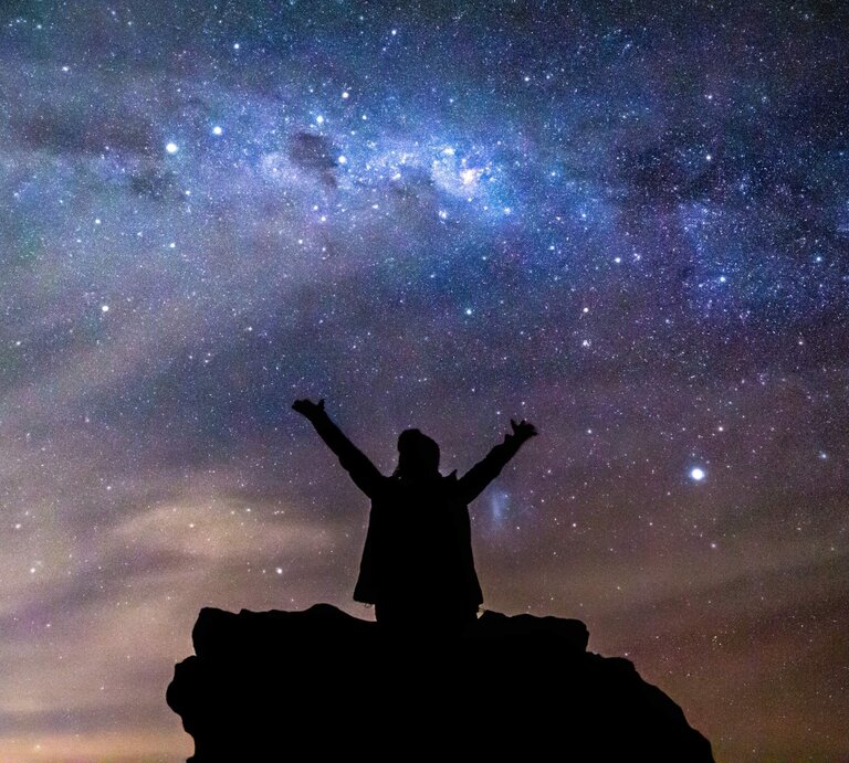 Eine Silhouette einer Person mit ausgestreckten Armen steht auf einem Felsen unter einem beeindruckenden Sternenhimmel. Die Milchstraße leuchtet hell über ihr, während der Himmel in tiefen Blau- und Violetttönen schimmert. Die Szene strahlt ein Gefühl von Freiheit, Staunen und Verbundenheit mit dem Universum aus. | © Lovleah via Canva.com