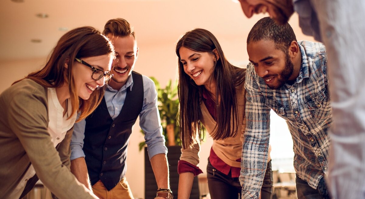 Eine Gruppe von fünf jungen Erwachsenen arbeitet gemeinsam an einem Tisch in einem modernen Büro. Sie sind in ein kreatives Projekt vertieft, lachen und zeigen Begeisterung. Eine Frau mit Brille und einem beigen Blazer zeigt auf ein Dokument oder eine Skizze, während die anderen aufmerksam zusehen. Die Gruppe ist vielfältig, mit verschiedenen Stilen und Hintergründen. Das warme Licht im Raum verleiht der Szene eine freundliche und motivierende Atmosphäre, die Zusammenarbeit und Teamgeist symbolisiert.
