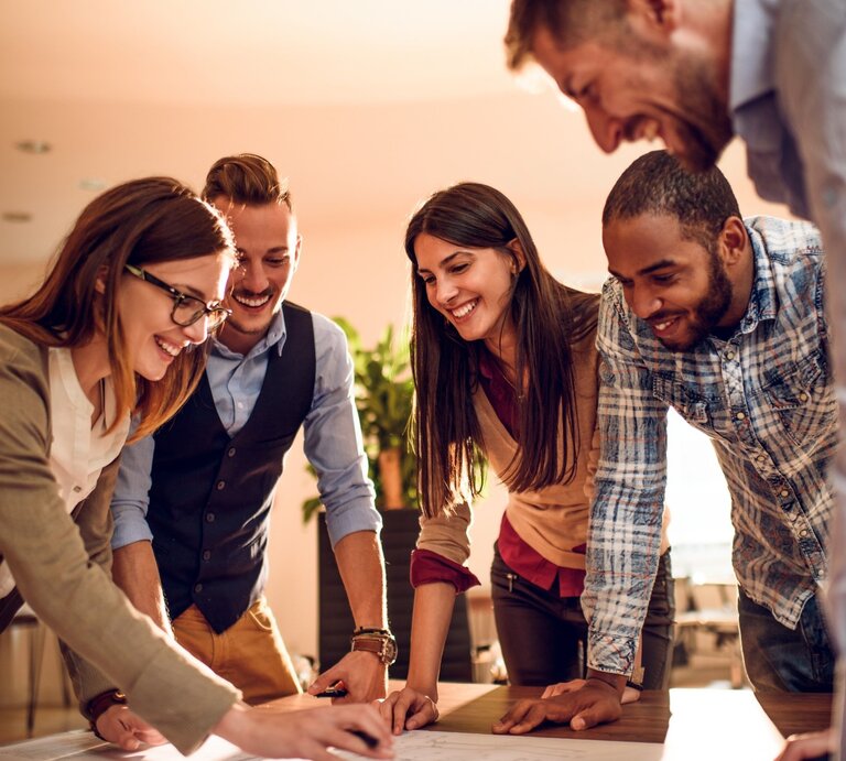 Eine Gruppe von fünf jungen Erwachsenen arbeitet gemeinsam an einem Tisch in einem modernen Büro. Sie sind in ein kreatives Projekt vertieft, lachen und zeigen Begeisterung. Eine Frau mit Brille und einem beigen Blazer zeigt auf ein Dokument oder eine Skizze, während die anderen aufmerksam zusehen. Die Gruppe ist vielfältig, mit verschiedenen Stilen und Hintergründen. Das warme Licht im Raum verleiht der Szene eine freundliche und motivierende Atmosphäre, die Zusammenarbeit und Teamgeist symbolisiert. | © Getty Images Signature/ bogdankosanovic via Canva.com