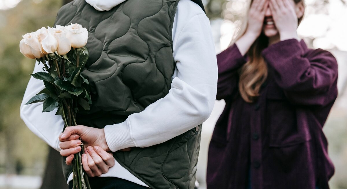 Eine Person in einer weißen Kapuzenjacke und grüner Weste hält hinter ihrem Rücken einen Strauß weißer Rosen, während eine andere Person mit langen Haaren und lila Jacke sich die Augen mit beiden Händen verdeckt und lächelnd auf die Überraschung wartet. Die Szene spielt sich in einem Park mit unscharfem Hintergrund aus Bäumen und Natur ab. Die Atmosphäre wirkt romantisch und verspielt.