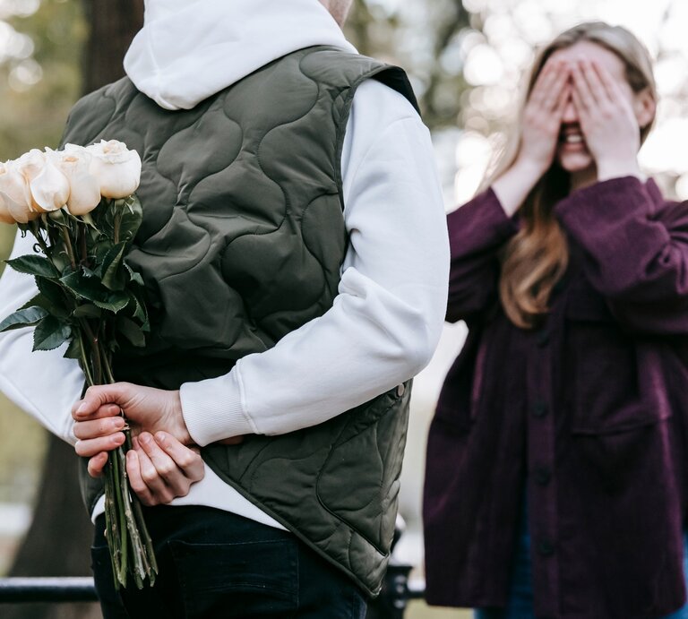 Eine Person in einer weißen Kapuzenjacke und grüner Weste hält hinter ihrem Rücken einen Strauß weißer Rosen, während eine andere Person mit langen Haaren und lila Jacke sich die Augen mit beiden Händen verdeckt und lächelnd auf die Überraschung wartet. Die Szene spielt sich in einem Park mit unscharfem Hintergrund aus Bäumen und Natur ab. Die Atmosphäre wirkt romantisch und verspielt. | © Pexels/ Katerina Holmes via Canva.com