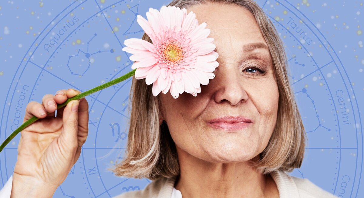 Eine ältere Frau mit schulterlangem, grauem Haar hält eine rosa Blume vor ihr Auge und lächelt sanft. Im Hintergrund ist ein astrologisches Rad mit Sternzeichen und Konstellationen auf einem blauen, sternenübersäten Himmel zu sehen. Das Bild vermittelt eine positive und harmonische Atmosphäre, die Astrologie und Lebensfreude verbindet.