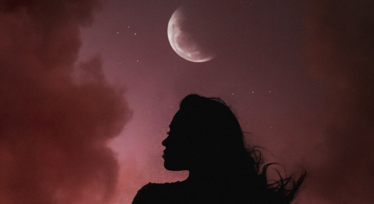 Silhouette einer Frau mit wehenden Haaren vor einem Nachthimmel mit rötlichen Wolken. Über ihr leuchtet ein zunehmender Halbmond, umgeben von kleinen Sternen. Die Szene wirkt mystisch und atmosphärisch.