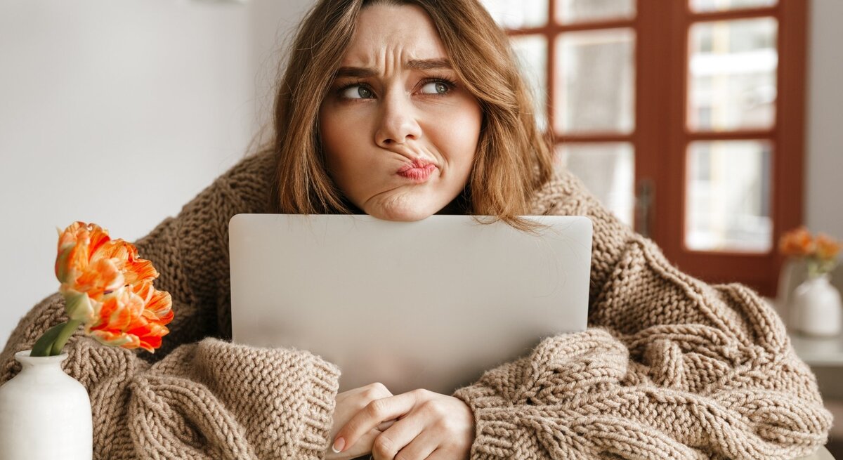 Eine nachdenkliche Frau stützt ihr Kinn auf einen Laptop, während sie einen skeptischen Gesichtsausdruck zeigt. Sie trägt einen dicken, beige gestrickten Pullover und sitzt an einem Tisch. Links von ihr steht eine weiße Vase mit einer orangefarbenen Blume. Im Hintergrund sind Fenster mit Holzrahmen zu sehen, durch die Tageslicht hereinfällt. Die Szene vermittelt eine gemütliche, aber nachdenkliche Stimmung.