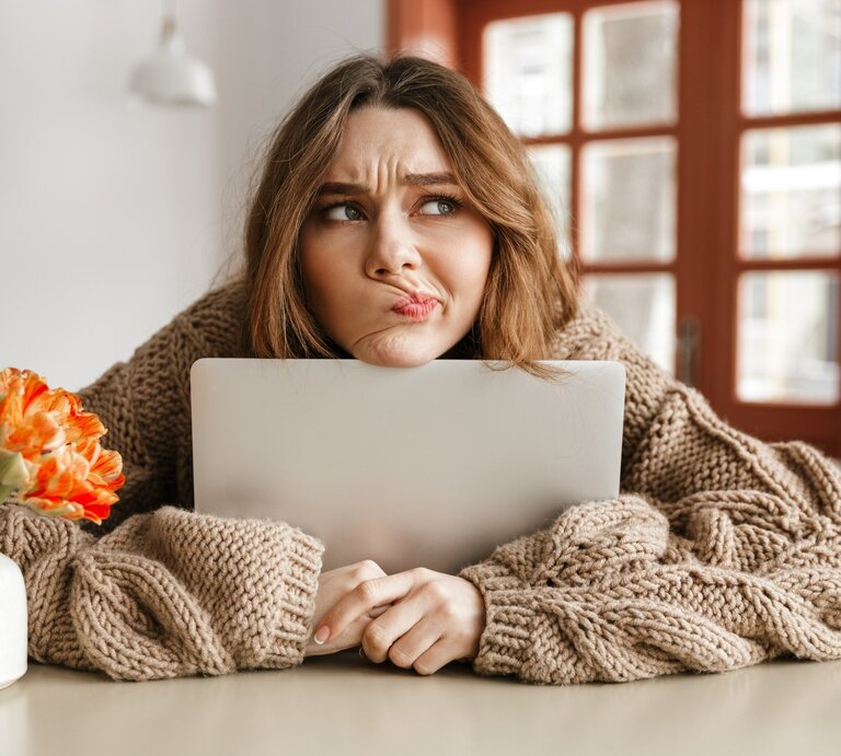 Eine nachdenkliche Frau stützt ihr Kinn auf einen Laptop, während sie einen skeptischen Gesichtsausdruck zeigt. Sie trägt einen dicken, beige gestrickten Pullover und sitzt an einem Tisch. Links von ihr steht eine weiße Vase mit einer orangefarbenen Blume. Im Hintergrund sind Fenster mit Holzrahmen zu sehen, durch die Tageslicht hereinfällt. Die Szene vermittelt eine gemütliche, aber nachdenkliche Stimmung. | © Dean Drobot via Canva.com 