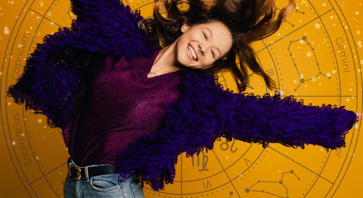 Eine fröhliche Frau mit langen, welligen Haaren springt lächelnd in die Luft. Sie trägt eine violette, flauschige Jacke und ein glitzerndes Oberteil, kombiniert mit einer Jeans. Der Hintergrund ist gelb mit einem astrologischen Tierkreis-Muster, das verschiedene Sternzeichen und Sternbilder zeigt.