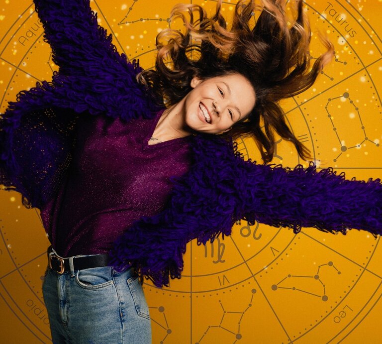 Eine fröhliche Frau mit langen, welligen Haaren springt lächelnd in die Luft. Sie trägt eine violette, flauschige Jacke und ein glitzerndes Oberteil, kombiniert mit einer Jeans. Der Hintergrund ist gelb mit einem astrologischen Tierkreis-Muster, das verschiedene Sternzeichen und Sternbilder zeigt. | © Getty Images Signature/ SolStock, Mas Water und WinWin.artlab via Canva.com [M]