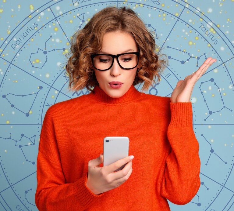  Eine junge Frau mit blonden, lockigen Haaren und einer schwarzen Brille hält ein Smartphone in der Hand und schaut überrascht oder nachdenklich darauf. Sie trägt einen leuchtend orangefarbenen Pullover. Im Hintergrund ist ein astrologisches Rad mit den Sternzeichen und funkelnden goldenen Details auf einem hellblauen Hintergrund zu sehen. Das Bild kombiniert moderne Technologie mit einem Hauch von Mystik und Astrologie und vermittelt eine dynamische, nachdenkliche Atmosphäre. | © Getty Images/ evgenyatamanenko, Mas Water und WinWin.artlab via Canva.com [M]
