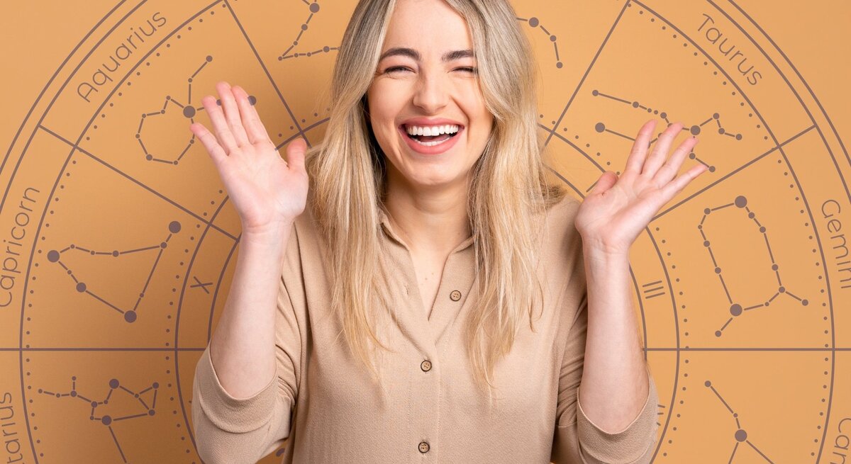 Eine fröhliche, blonde Frau mit langen Haaren lächelt breit und hebt beide Hände freudig in die Luft. Sie trägt ein beigefarbenes Oberteil. Im Hintergrund ist ein großes astrologisches Rad mit den Symbolen und Namen der Sternzeichen wie Aries, Taurus, Gemini und Pisces zu sehen. Die warmen, erdigen Farbtöne des Bildes vermitteln eine positive und optimistische Stimmung.