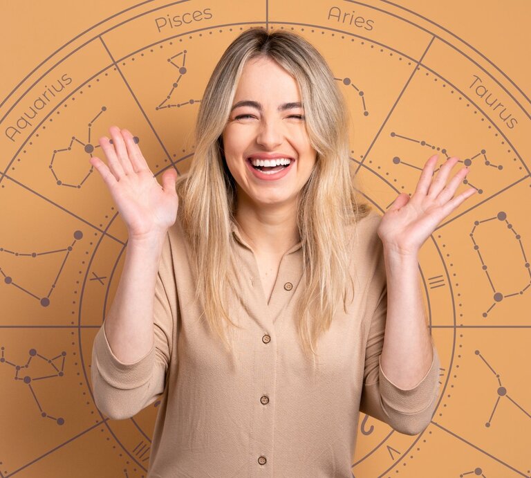 Eine fröhliche, blonde Frau mit langen Haaren lächelt breit und hebt beide Hände freudig in die Luft. Sie trägt ein beigefarbenes Oberteil. Im Hintergrund ist ein großes astrologisches Rad mit den Symbolen und Namen der Sternzeichen wie Aries, Taurus, Gemini und Pisces zu sehen. Die warmen, erdigen Farbtöne des Bildes vermitteln eine positive und optimistische Stimmung. | © Getty Images/ Vergani_Fotografia und WinWIn.artlab via Canva.com [M]