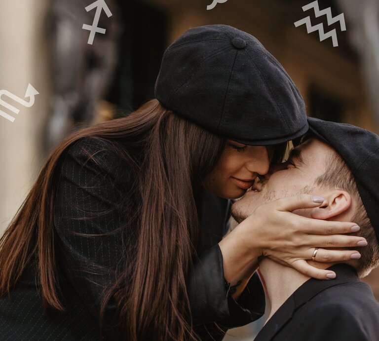 Ein verliebtes Paar in dunkler Kleidung und passenden schwarzen Mützen, das sich zärtlich küsst. Die Frau hält das Gesicht des Mannes liebevoll mit beiden Händen. Im Hintergrund sind dezent astrologische Sternzeichensymbole zu sehen. | © Pexels/ Zhanna Fort und Chikovnaya via Canva.com [M]