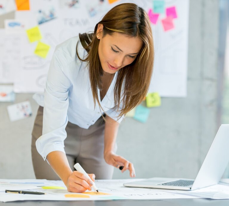 Junge Frau in Business-Kleidung arbeitet konzentriert an einem Schreibtisch, schreibt Notizen auf ein großes Papier und hat einen Laptop sowie bunte Haftnotizen im Hintergrund. | © Getty Images Signature/ andresr via Canva.com