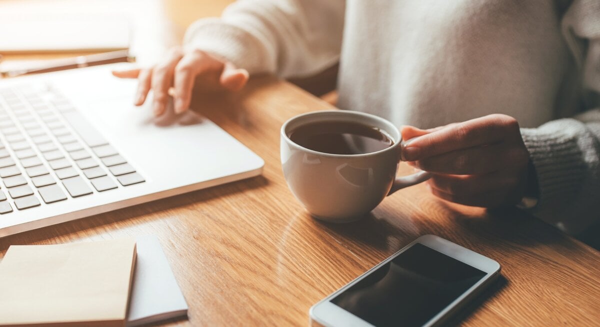 Eine Person sitzt an einem Holztisch und arbeitet an einem Laptop. In der rechten Hand hält sie eine weiße Tasse mit heißem Kaffee, während die linke Hand die Tastatur bedient. Neben dem Laptop liegen Notizzettel und ein Stift. Auf dem Tisch befindet sich außerdem ein Smartphone mit schwarzem Bildschirm. Durch ein Fenster im Hintergrund fällt warmes, natürliches Licht und erzeugt eine gemütliche Atmosphäre.