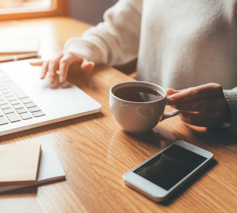 Eine Person sitzt an einem Holztisch und arbeitet an einem Laptop. In der rechten Hand hält sie eine weiße Tasse mit heißem Kaffee, während die linke Hand die Tastatur bedient. Neben dem Laptop liegen Notizzettel und ein Stift. Auf dem Tisch befindet sich außerdem ein Smartphone mit schwarzem Bildschirm. Durch ein Fenster im Hintergrund fällt warmes, natürliches Licht und erzeugt eine gemütliche Atmosphäre. | © annakimphotos via Canva.com