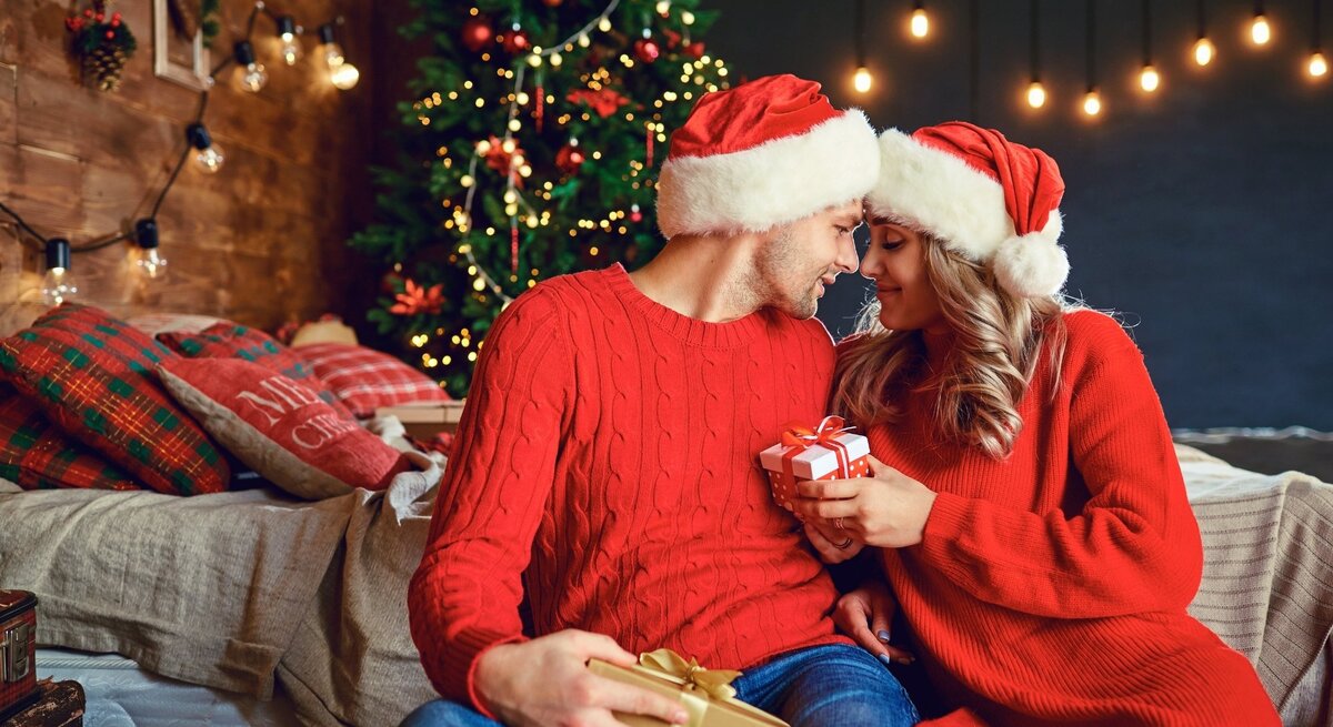 Ein verliebtes Paar in roten Weihnachtsoutfits und Weihnachtsmützen sitzt vor einem festlich geschmückten Weihnachtsbaum. Sie halten sich gegenseitig Geschenke und lächeln sich zärtlich an, während im Hintergrund eine gemütliche, weihnachtliche Szenerie mit Lichterketten und dekorierten Kissen zu sehen ist.